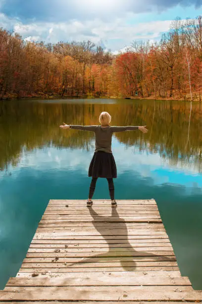Woman standing on the edge of wooden dock, with her arms raised up, looking at water and colorful autumnal forest