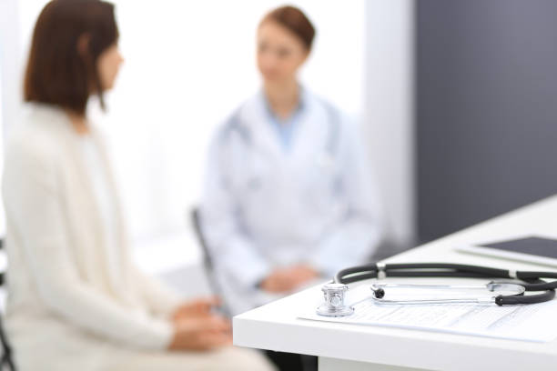istock Stethoscope, clipboard with medical form lying on hospital reception desk with laptop computer and busy doctor and patient communicating at the background. Medical tools at doctor working table.Medicine concept 1066088338