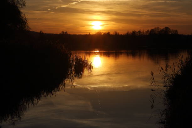 istock Dramatic sunset over a lake and a stream flowing into it. Orange sunlight reflecting on water surface, deep shadows of forest and and water plants. 1066113190