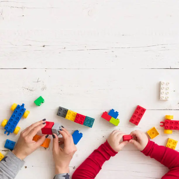 Photo of Closeup of young woman's and child's hands playing with toys and plastic bricks on white wooden table background