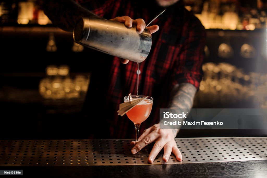 Barman pouring fresh Paper Plane cocktail into a decorated glass Barman pouring fresh Paper Plane cocktail from shaker into a decorated glass on the bar counter Cocktail Stock Photo