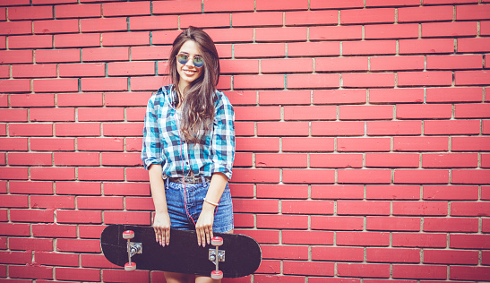 Cute girl in checkered shirt standing in front of red brick wall with her skateboard