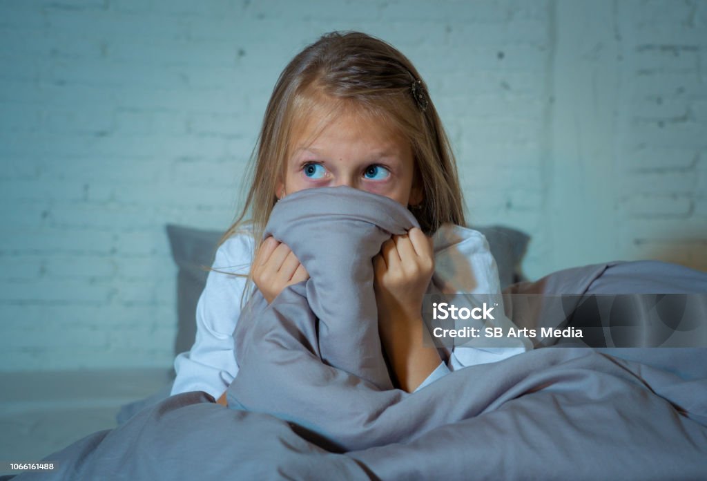 Scared little girl staying sleepless hiding behind the duvet looking horrified in the dark having childhood nightmares in child imagination Sleeping disorders Stress Depression and Insomnia concept. Child Stock Photo