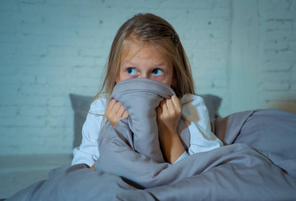 Scared little girl staying sleepless hiding behind the duvet looking horrified in the dark having childhood nightmares in child imagination Sleeping disorders Stress Depression and Insomnia concept. Scared little girl staying sleepless hiding behind the duvet looking horrified in the dark having childhood nightmares in child imagination Sleeping disorders Stress Depression and Insomnia concept. fearfull stock pictures, royalty-free photos & images