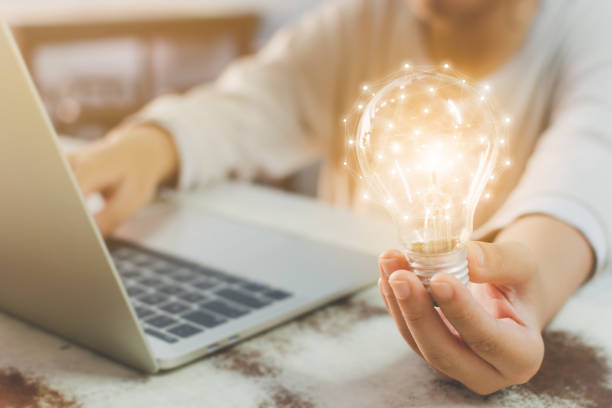 рука женщины держа лампочку и используя laptop на деревянном столе. концепция новой идеи с инновациями и творчеством - hardy bulbs стоковые фото и изображения