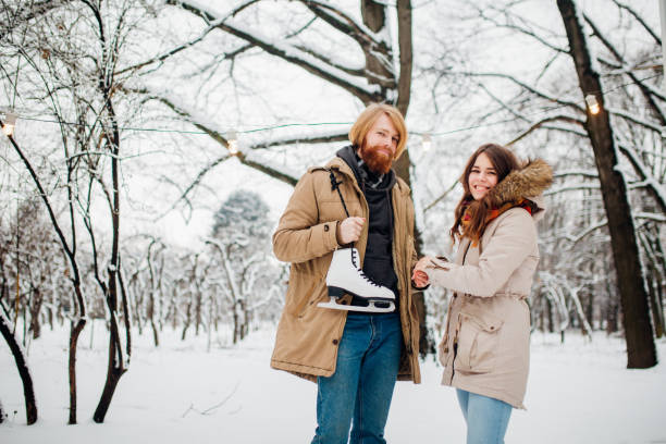 winter and date. young couple, lovers man and woman in winter on a background of snow and forest holding hands and smiling. a guy with long hair and a beard is holding skates and looking at the girl - long hair red hair women men imagens e fotografias de stock