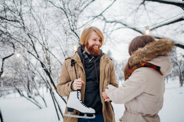 winter and date. young couple, lovers man and woman in winter on a background of snow and forest holding hands and smiling. a guy with long hair and a beard is holding skates and looking at the girl - long hair red hair women men imagens e fotografias de stock