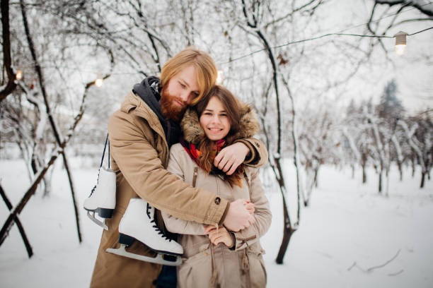 winter and date. young couple in love with man and woman in the winter against background of snow covered trees in park are embrace. a guy with long hair and beard keeps skates and loves girlfriend - long hair red hair women men imagens e fotografias de stock