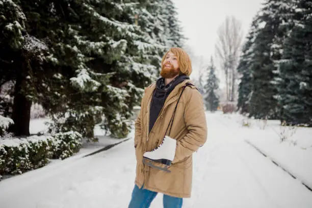 Winter sports theme. Portrait. Handsome young Caucasian guy with long hair and red beard. Model poses in winter snow park against background of the forest on the shoulder of a pair of leather skates.