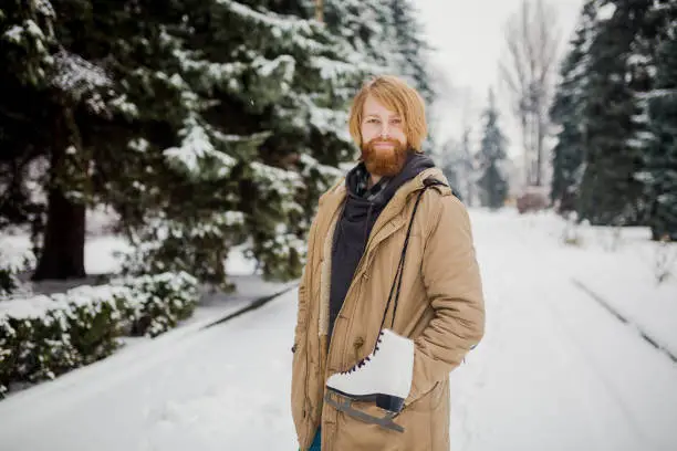 Winter sports theme. Portrait. Handsome young Caucasian guy with long hair and red beard. Model poses in winter snow park against background of the forest on the shoulder of a pair of leather skates.