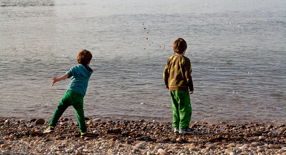 Two young boys playing on a pebble beach, chasing bubbles to pop and throwing stones in the water. Colour image.