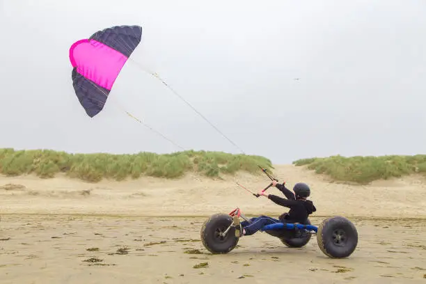 Dutch teenage boy driving kite buggy with kite flyer on beach. This photo from my youngest son was taken during our vacation in the province Zeeland in the Netherlands. This area is located at the coast with beach and dunes. The teenage boy was transported by a kite buggy. He was easily transported along the coast by the wind. Steering is done through the legs by turning the front wheel.