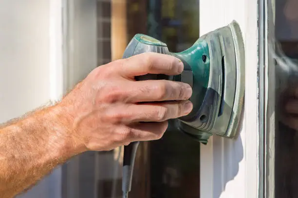 Hand of painter sanding window frame with electrical sander. This male hand is holding a tool to sand the wood of this house. This is necessary to prepare the construction for painting.