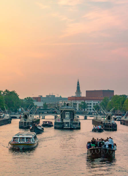 Multiple boats headed towards some locks on the canals of Amsterdam.  Beautiful sunset sky in the distance of orange, punk and blue.  Clouds in the sky.