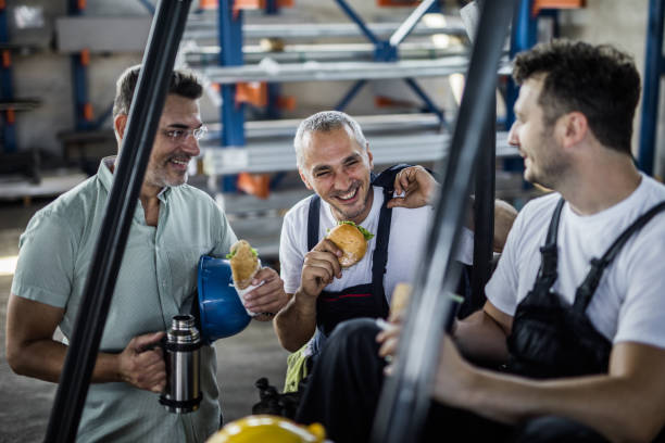 Happy manual workers and their manager talking on a lunch break in a factory. Group of workers communicating while eating sandwiches on a lunch break in industrial building. construction lunch break stock pictures, royalty-free photos & images