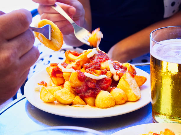 Patatas Bravas, fried potatoes with spicy sauce, one of the most common typical Spanish tapas."n People taste a portion of Patatas Bravas over a metallic table. Fried potatoes topped with spicy sauce, also know as Patatas a la Brava or Papas Bravas, one of the most common typical Spanish tapas. patatas bravas stock pictures, royalty-free photos & images