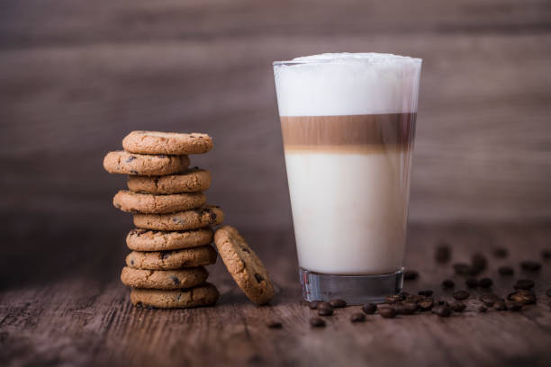 Chocolate chip cookies and a layered cafe latte coffee drink Cookies and coffee - a high transparent glass filled with a layered cafe latte on a wooden table, next to a pile of chocolate chip cookies. Some roasted coffee beans are spread out on the table. Horizontal photo. macchiato stock pictures, royalty-free photos & images