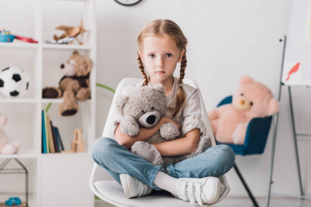 istock lonely little child sitting on chair and embracing her teddy bear while looking at camera 1066370570