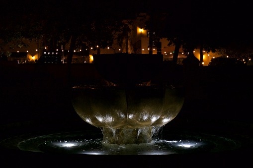 This photo was taken at Libbey Park in Ojai, Ventura County, California, USA, on July 15, 2018, at nighttime. We see the Fountain, and behind it, in the background, we can see the lights of the shops and restaurants adjacent to the east border of the park.