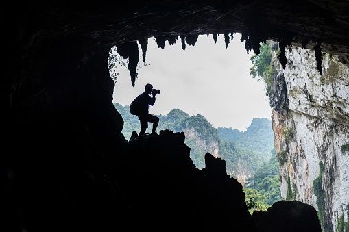 Eco-tourism - silhouette of solo tourist exploring cave and capturing view with camera, limestone karst cliff in background.