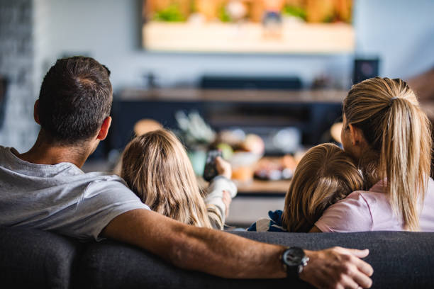 Rear view of a family watching TV on sofa at home. Back view of a relaxed family watching TV on sofa in the living room. television set stock pictures, royalty-free photos & images