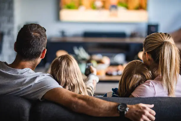 Photo of Rear view of a family watching TV on sofa at home.