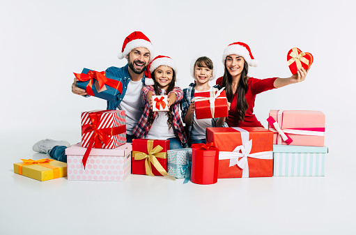 Young happy family in Santa hats with pile of christmas presents show it into the camera isolated on white background. New Year holidays