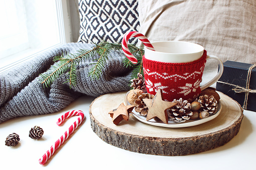 Winter morning breakfast still life scene. Cup of coffee, tea with candy canes standing near window on wooden cut board. Decorated by pine cones, hazelnuts and wooden stars. Christmas concept.