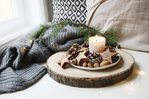 Winter festive still life scene. Burning candle decorated by wooden stars, hazelnuts and pine cones standing near window on wooden cut board. Glittering Christmas lights, fir branch on wool plaid.