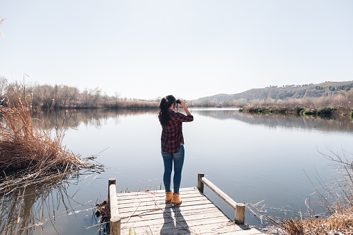 Young brunette woman on a lake's jetty with binoculars and plaid shirt