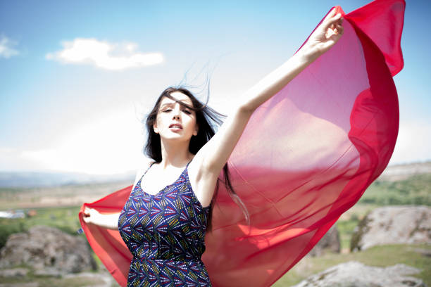 A woman in a red dress stands in a lush field of red poppies (Papaver rhoeas). She wears a black and red patterned necklace and a sheer black scarf draped over her shoulders. Her expression is serene, eyes closed, as if enjoying the moment. The sky is partly cloudy, and trees line the background, adding to the sense of a tranquil, natural setting.