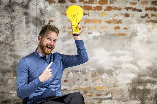 Happy redhead businessman aiming at light bulb placard and looking at camera. Copy space.