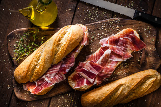 Preparing iberico ham sandwich, Spanish sandwich of iberian ham Typical spanish food concepts: High angle view of a rustic wooden table with two delicious Iberico ham sandwich also called in Spain as Bocadillo de Jamón Iberico. Predominant color is brown. Low key DSRL studio photo taken with Canon EOS 5D Mk II and Canon EF 100mm f/2.8L Macro IS USM. appetizer stock pictures, royalty-free photos & images