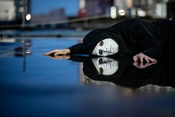 reflexão do homem de capuz misterioso no sentimento de máscara branca deprimido e decepcionado deitado no chão molhado, depois da chuva. conceitos de tristeza, depressão e transtorno bipolar - falling suicide men built structure - fotografias e filmes do acervo