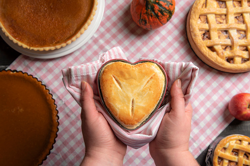 Woman holding a raspberry pie, in a heart-shaped tray,  above a kitchen table full of traditional fall pies, apple pies, and pumpkin pies.