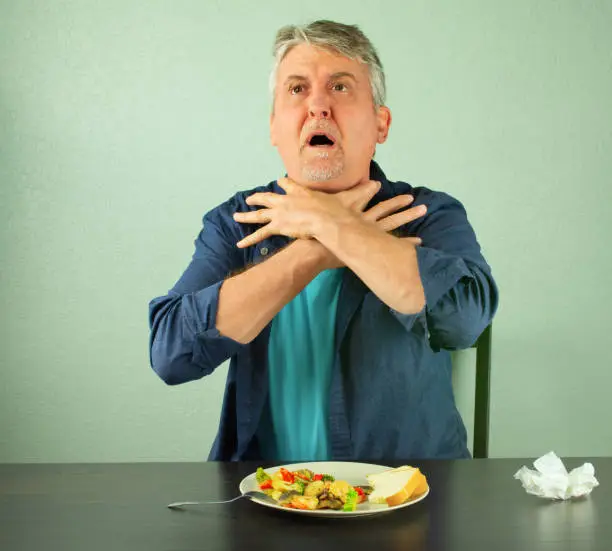 A man is panicking and is in trouble as he chokes on food and is making the official international sign for "I'm choking" by placing his hands around his neck with outstretched fingers.