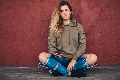 Portrait of a young woman dressed in a hoodie and ripped jeans leaning on a wall while sitting on a skateboard at bridge footway.
