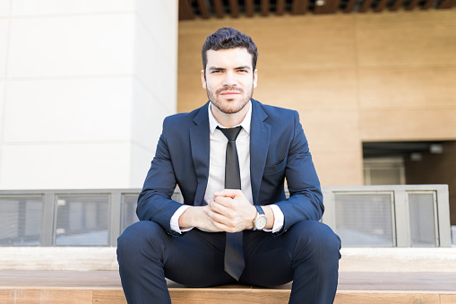 Portrait of attractive sales manager wearing elegant suit while sitting on bench in city