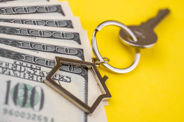 The concept of buying a private house or apartment. Mortgage registration. The accumulation of money for real estate. Metal key chain in the shape of a house with a metal key lying on the pack of hundred US dollars on a yellow background. Close up. el concepto de comprar una casa o un apartamento. inscripción de la hipoteca. la acumulación de dinero para bienes raíces. cadena dominante del metal en forma de una casa con una llave de metal en el paquete de cientos de millones de dólares en un fond