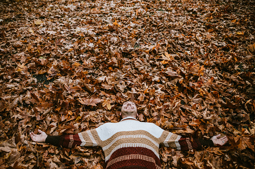 An adult man smiles, having fun laying on a leaf colored field that matches the colors of his knit sweater.  Horizontal image with copy space.