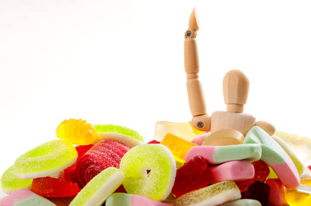 Unhealthy eating habits, November 14 is diabetes awareness day and bad diet concept with a puppet drowning in sugary sweets and junk food isolated on white background and copy space for text hábitos alimenticios poco saludables, 14 de noviembre es día de concientización de diabetes y la insulina resistencia y el concepto de la mala dieta con una marioneta ahogado en azucaradas dulces y comida chatarra, aislado en fondo blanco y copyspace pa