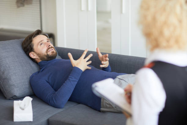 istock Depressed handsome young bearded man in turtleneck sweater suffering from personal crisis lying on gray sofa and gesturing hands while sharing his mental problems, he visiting psychologist 1066705472