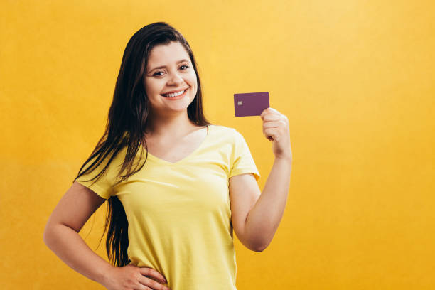 retrato de uma jovem feliz, mostrando o cartão de crédito plástico isolado sobre fundo amarelo - showing buying paying clipping path - fotografias e filmes do acervo