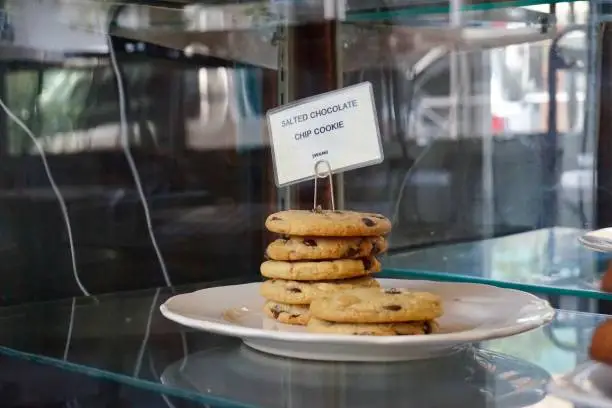 Chocolate chip cookies stacked on a plate, with a little label on top. Cookies are inside a glass display case in a coffee shop.