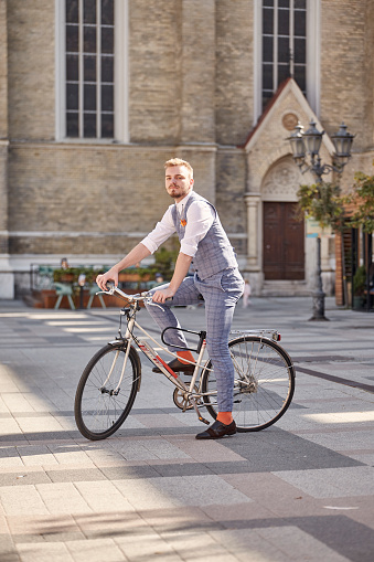 one young man, 20-29 years old, wearing hipster suit, smart casual,  posing on old city bike, outdoors on old European city square (out of focus).