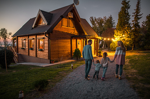 Rear view of happy family holding hands and walking by their chalet in the evening.