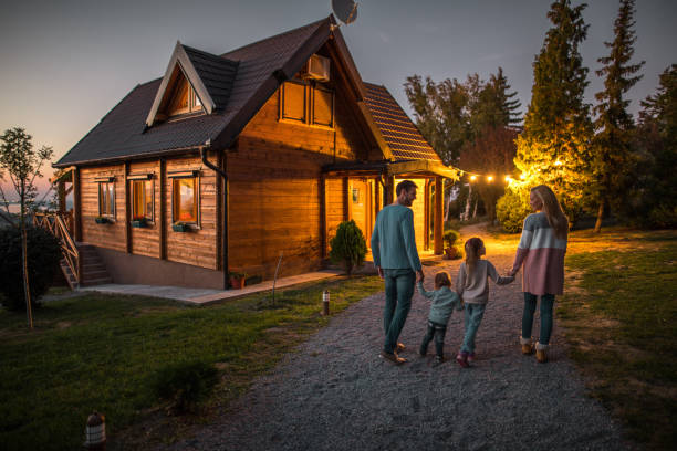 Rear view of happy family holding hands and walking by their chalet in the evening. Back view of young smiling parents holding hands with their children and walking in nature in the evening. chalet stock pictures, royalty-free photos & images