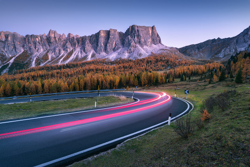 Blurred car headlights on winding road in mountains at sunset in autumn. Spectacular landscape with asphalt road, light trails, forest, rocks and purple sky at night in fall. Car driving on roadway