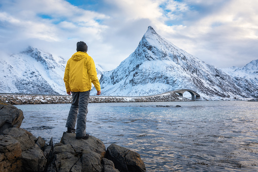 Sporty man in yellow jacket standing in the stone on seacoast against snowy mountains and cloudy sky at sunset in winter. Landscape with man, water, Fredvang bridge in Lofoten Islands, Norway.
