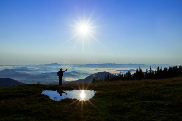istock High in the mountains above the clouds, a young man is looking for a cellular connection. Highly raising the phone, the sun looks into the camera with rays and is reflected in a puddle 1066926448